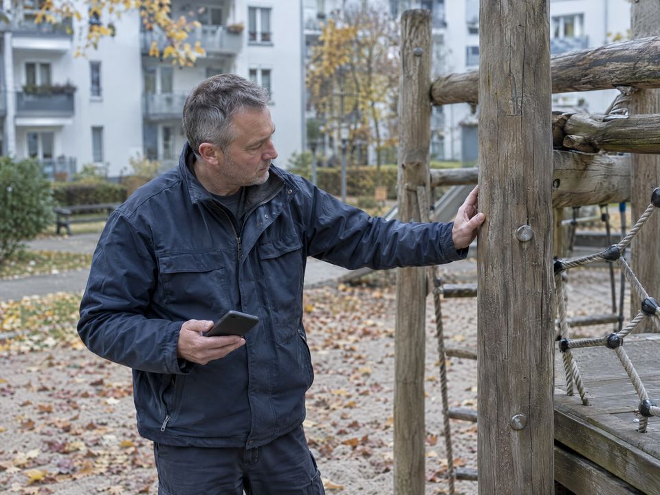 KI-Symbolbild: Ein Hausmeister prüft das Klettergerüst auf dem Spielplatz einer Wohnanlage, das Smartphone in der Hand.