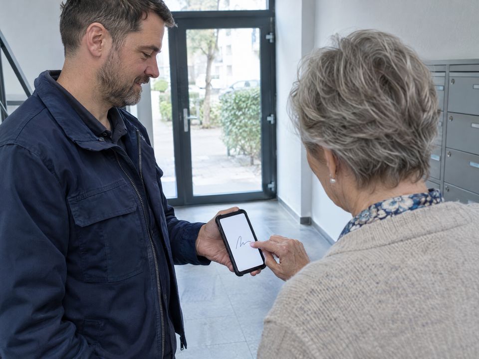 KI-Symbolbild: Im Hauseingang unterschreibt eine Frau mit dem Finger auf dem Smartphone, das ihr der Hausmeister hinhält.