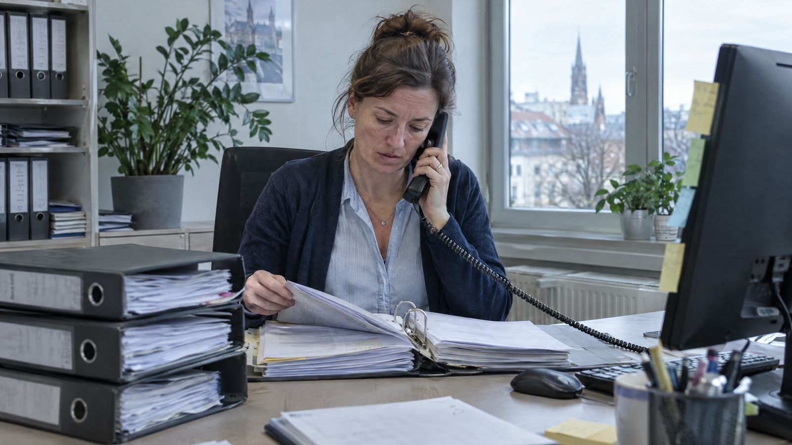KI-Symbolbild: Eine Hausverwalterin telefoniert an ihrem Schreibtisch und blättert dabei in einem Ordner, daneben stapeln sich weitere Ordner und Haftnotizen.