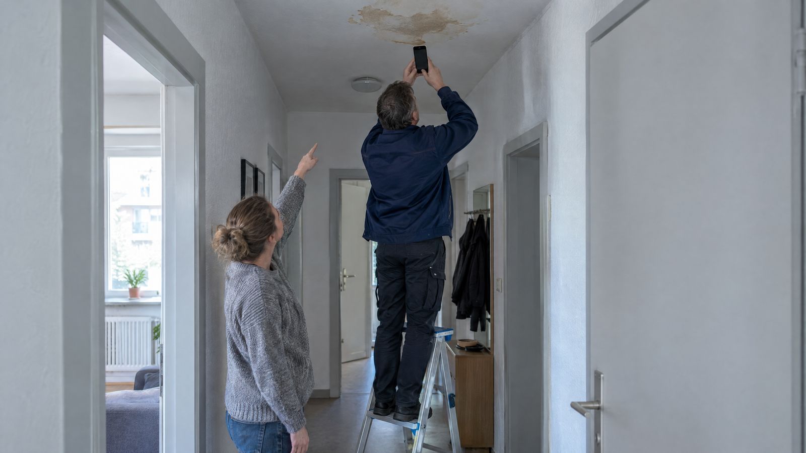 KI-Symbolbild: Ein Hausmeister fotografiert von der Leiter aus einen Wasserfleck an der Flurdecke, die Mieterin zeigt auf die Stelle.